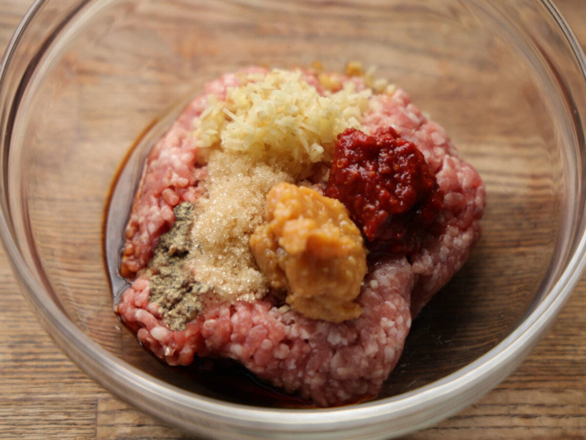 透明なボウルに豚ひき肉、みじん切りしょうが、砂糖、味噌、豆板醤などの調味料が入っている。 透明なボウルに豚ひき肉、みじん切りしょうが、砂糖、味噌、豆板醤などの調味料が入っている。