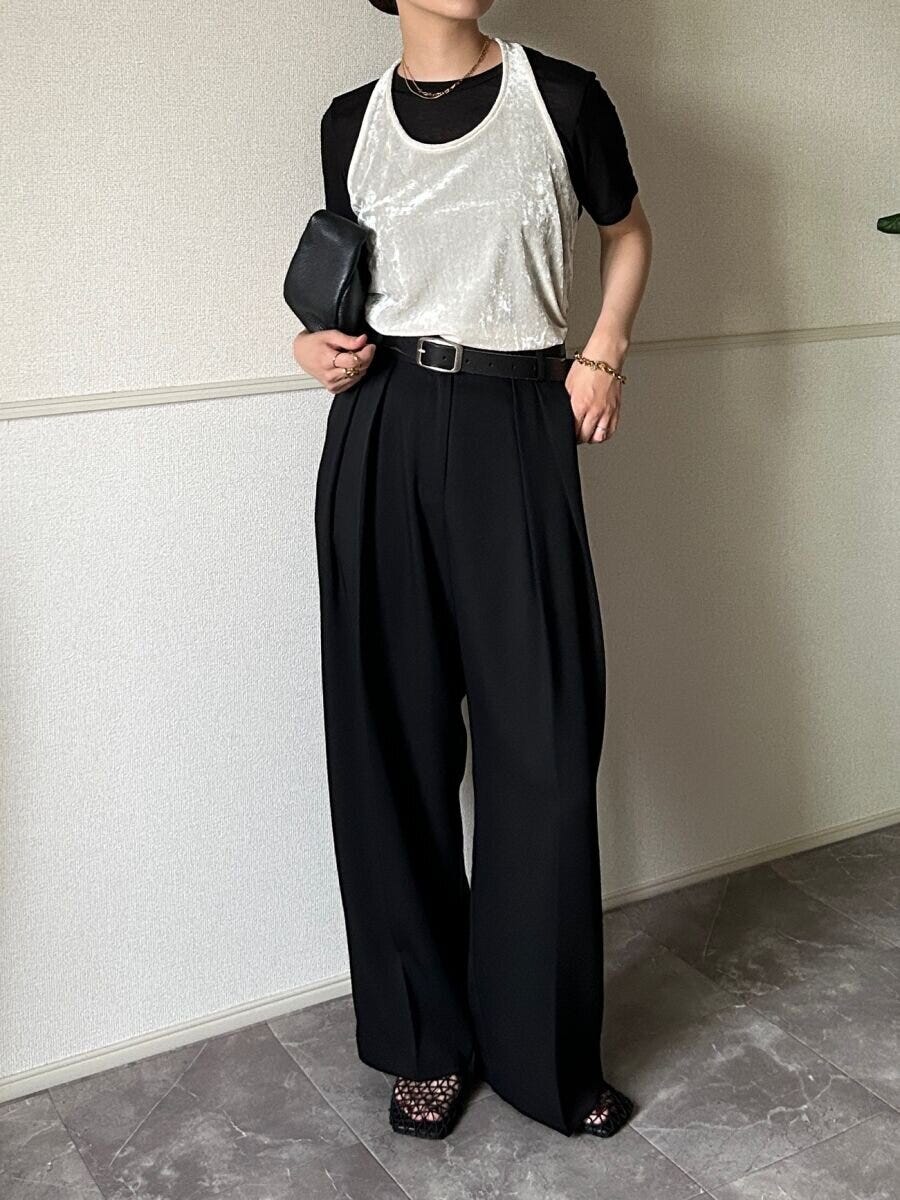 黒いシアーTシャツの上に白いベロア調タンクトップをレイヤードし黒いワイドパンツを着用した女性が立っている