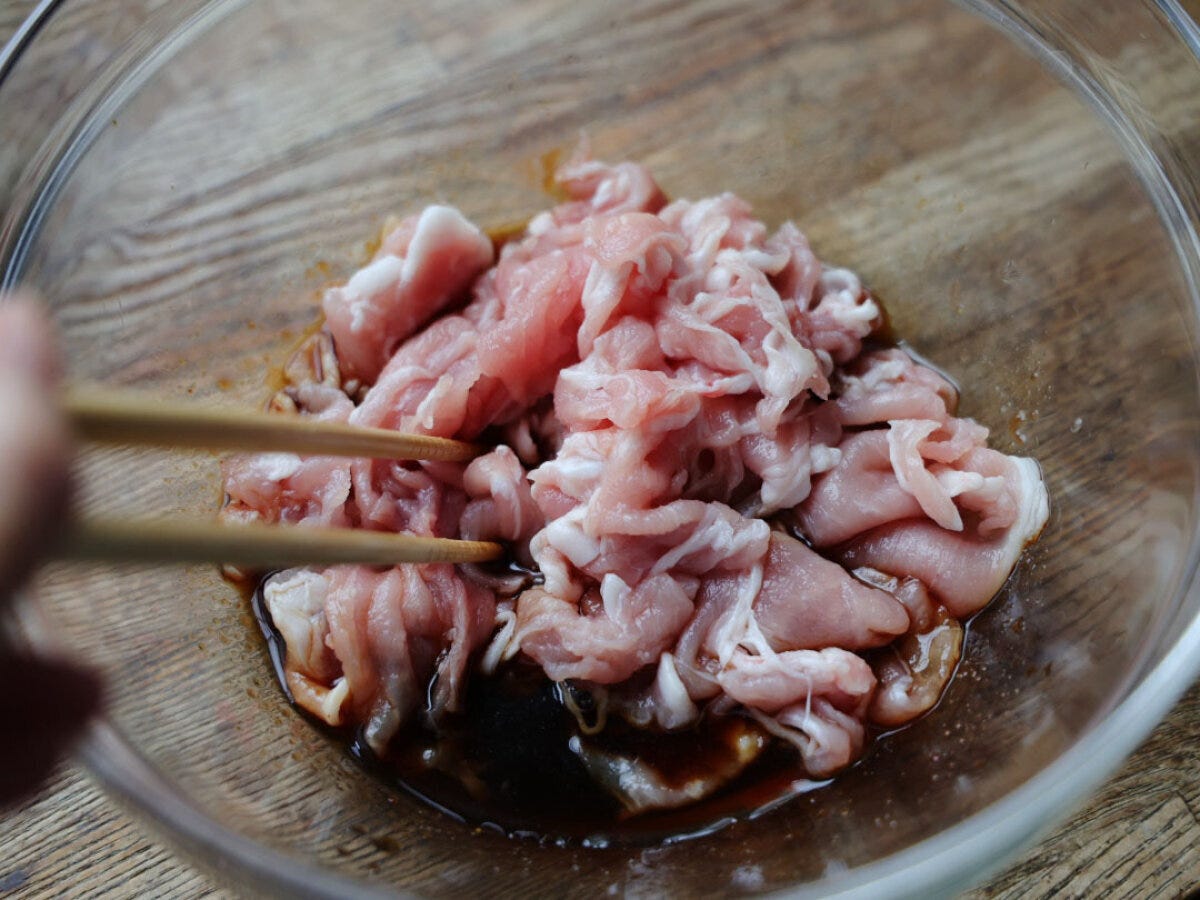 透明なボウルに入った調味料に薄切り豚肉を加え、箸で和えている