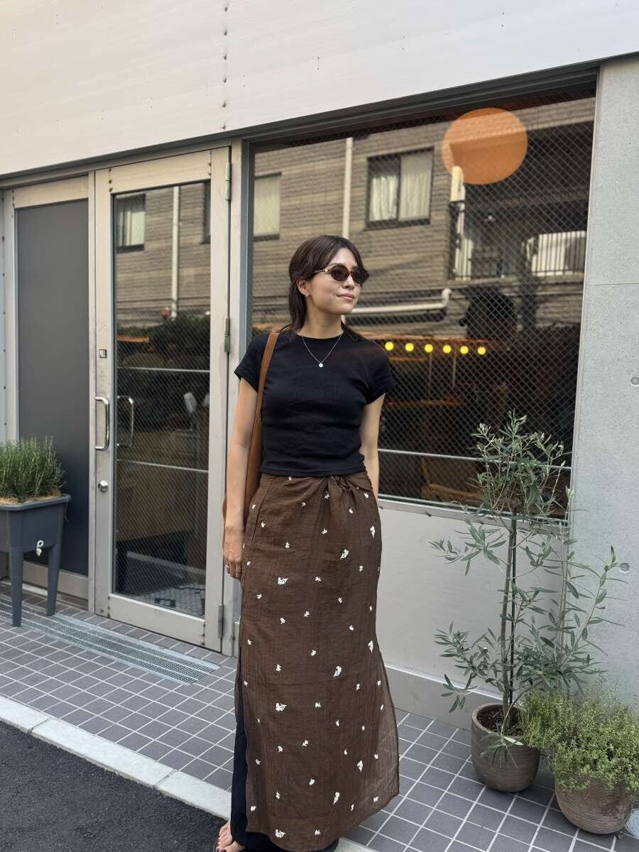 黒いミニTとブラウンのロングスカートを着用した女性が、店の前で立っている