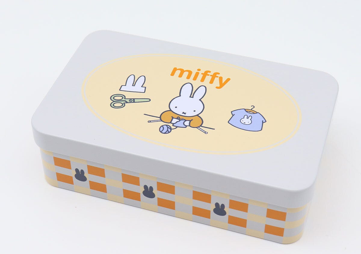 ミッフィーが描かれた白い缶ケース 白い長方形の缶ケースのふた、中央の黄色い楕円フレーム内にハサミと編み物するミッフィーとミッフィーTシャツが描かれ、側面はオレンジと水色のチェック柄