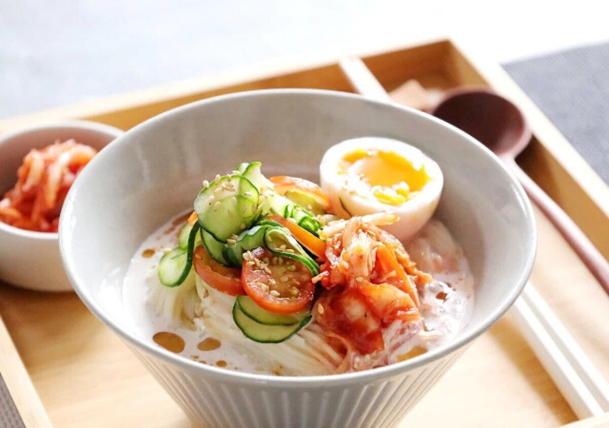 白い器に盛ったそうめんと豆乳スープのコングクスに、きゅうり・プチトマト・キムチ・半熟卵をのせ白ごまをふった写真