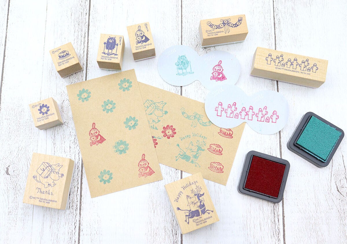 ムーミンの木製スタンプ10個と2色のインクパッド、スタンプを押したクラフト紙やカードが木製のテーブルに広げられている。