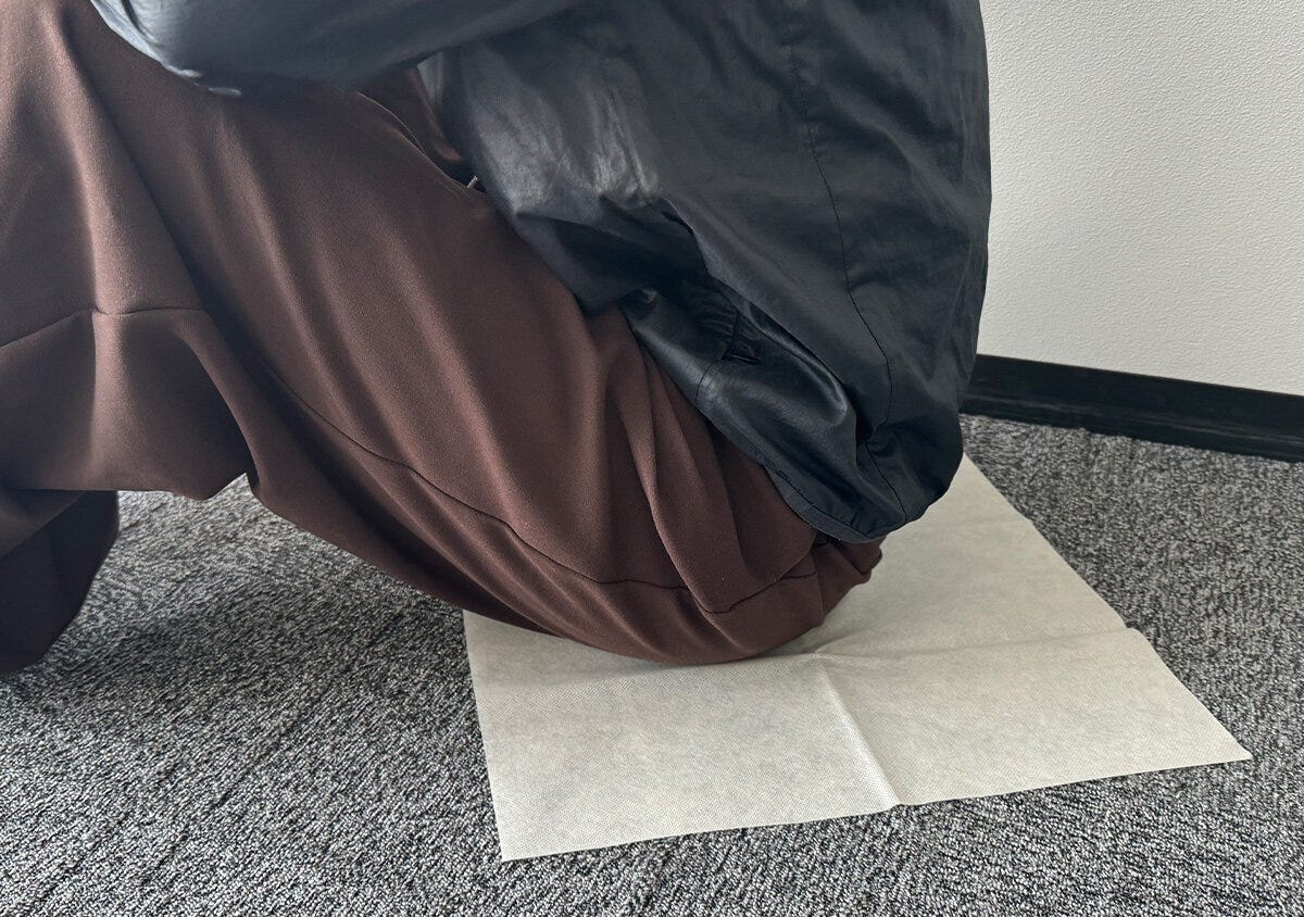 室内で黒い上着と茶色いパンツの人がレジャーシートに座っている様子