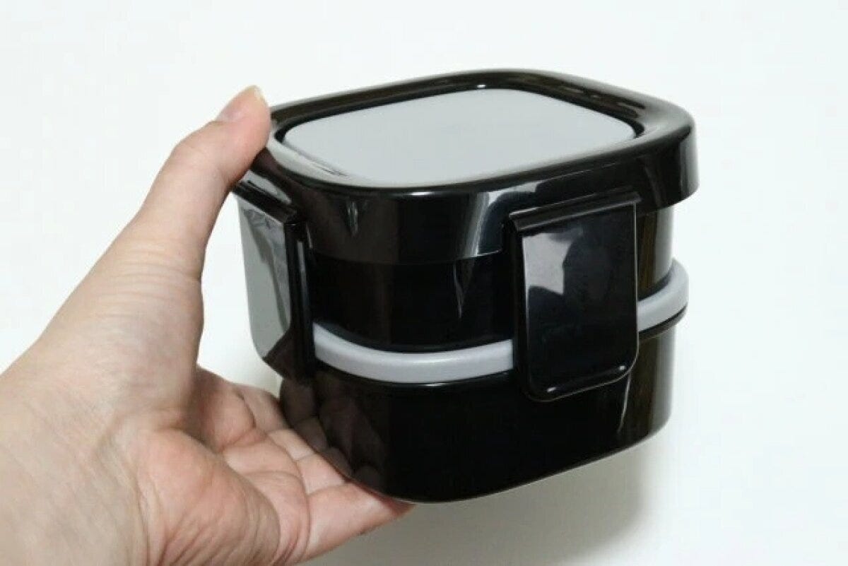 手で持たれた黒い二段弁当箱。ロック蓋が閉じた状態 手で持たれた黒い二段弁当箱。ロック蓋が閉じた状態