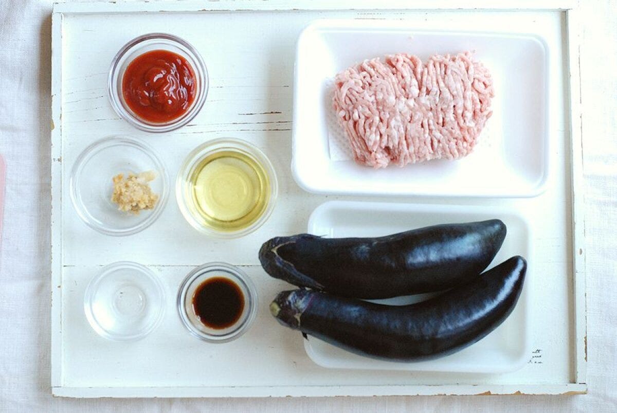 白いトレイの上に並べられた、なす、豚ひき肉、調味料（ケチャップ、油、味噌、酒、醤油、にんにく、砂糖）の材料