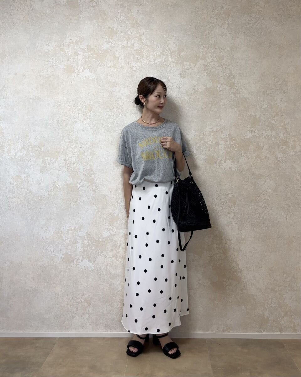白のドット柄スカートとグレーのロゴTシャツを着た女性が斜めを向いて立っている