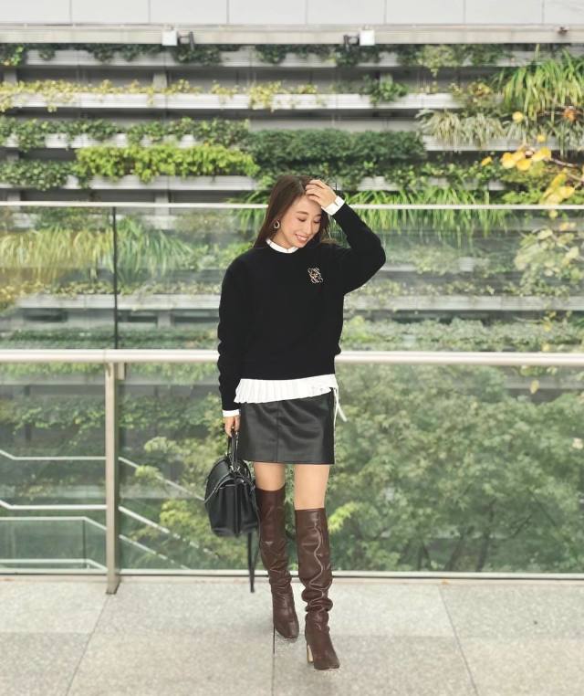 白のプリーツブラウスに黒のクルーネックニットを合わせ、黒のレザーミニスカートを穿き、ブラウンのレザーニーハイブーツと黒のハンドバッグをコーディネート