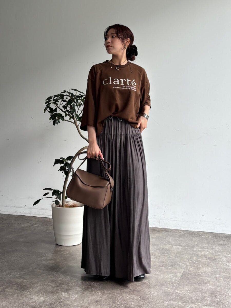 ブラウンのロゴTシャツとグレーのギャザースカートを着用し、ブラウンのバッグを持つ女性