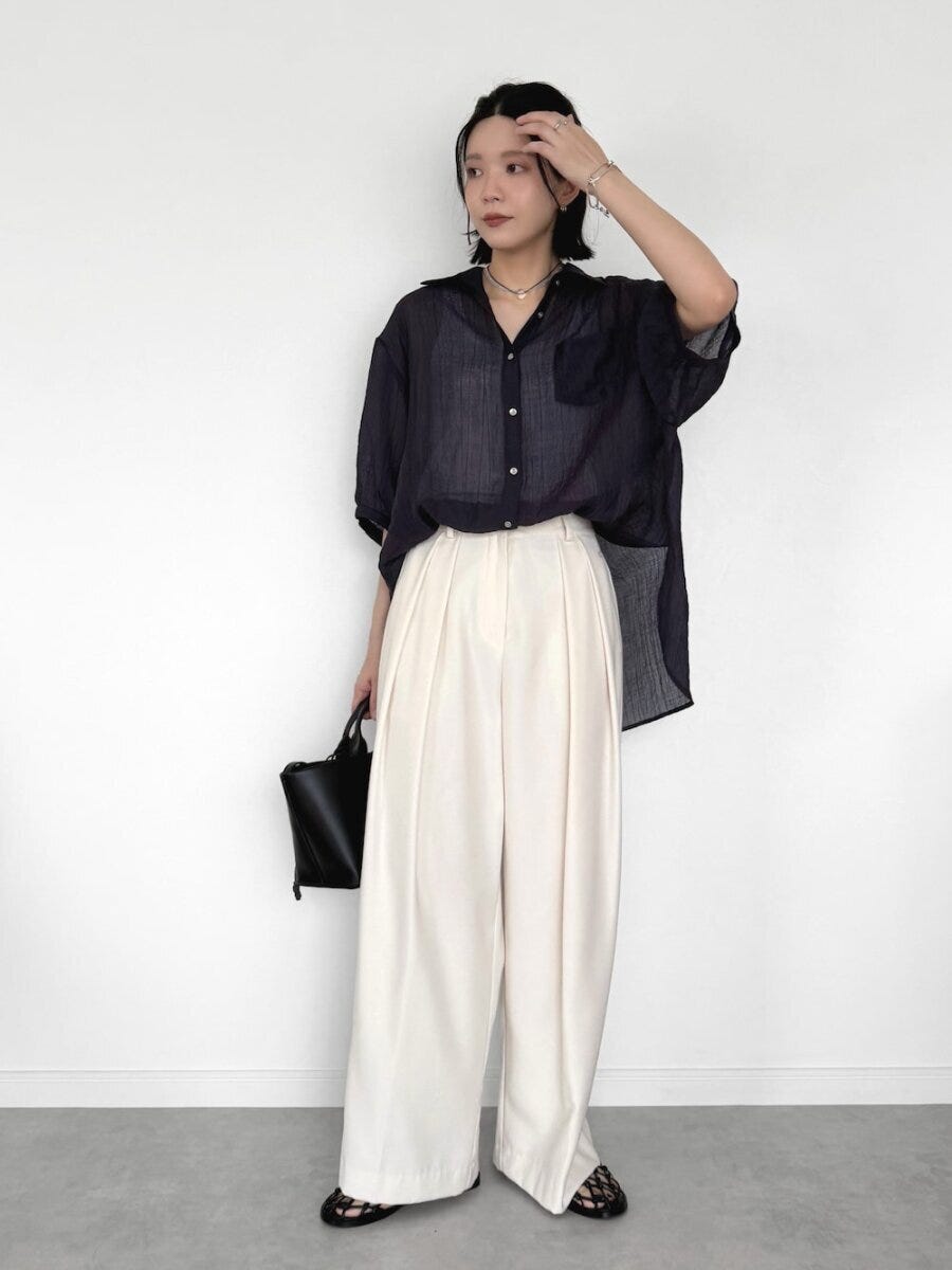 黒のシアーシャツと白のタックワイドパンツを着て髪に手を添える女性