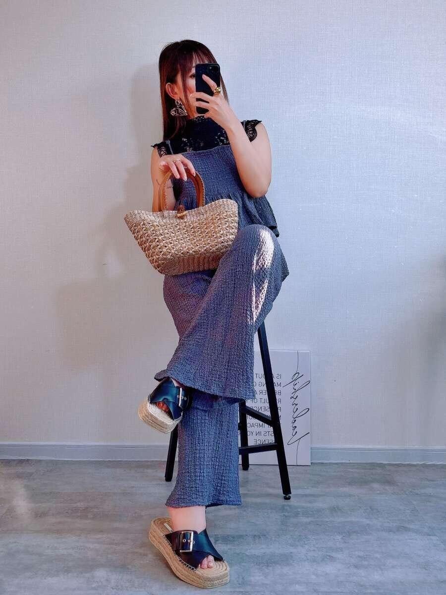 黒レースブラウスを着用した女性がグレーのオールインワンを着て椅子に座りかごバッグを持っている