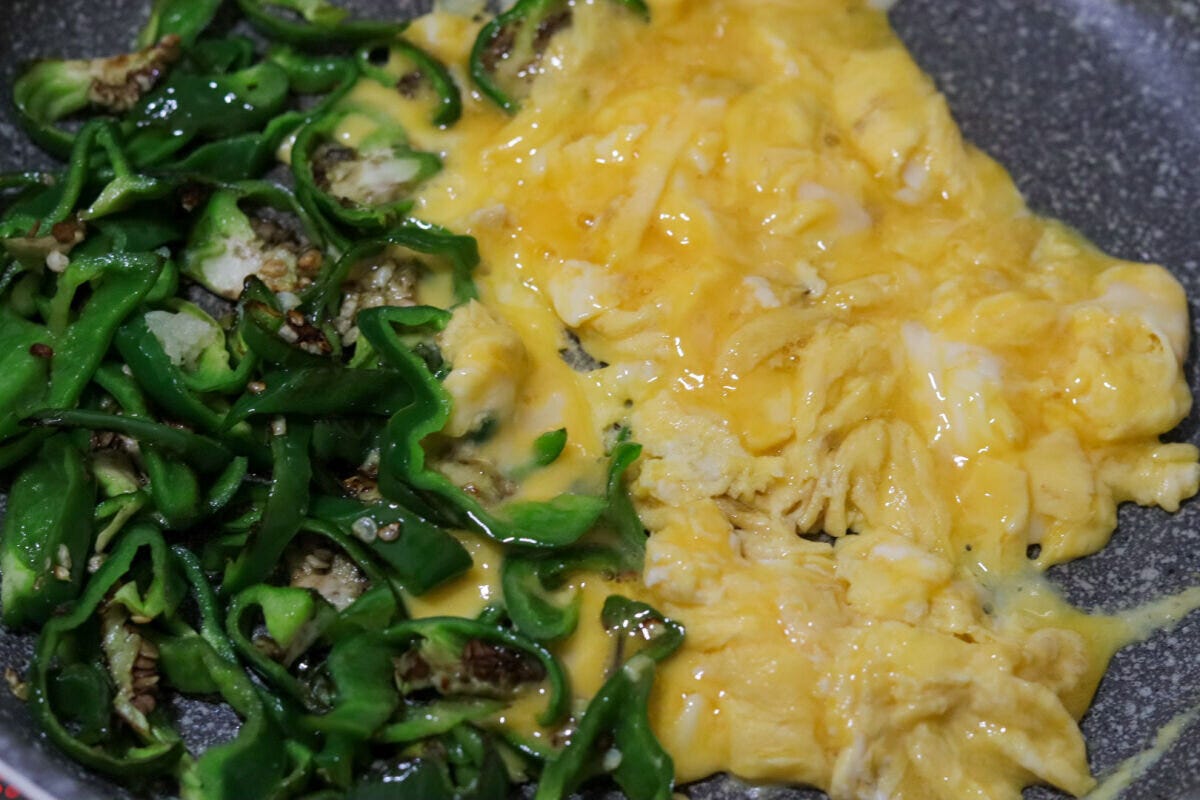 フライパンの片側に寄せられた焼きピーマンと、空いたスペースで半熟状に炒められている卵