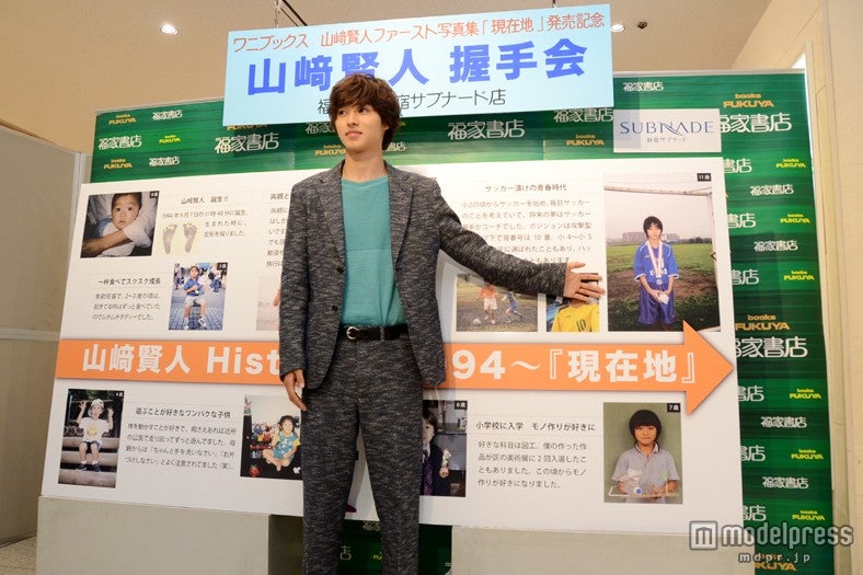 生い立ちを振り返る年表パネルをバックにポーズ