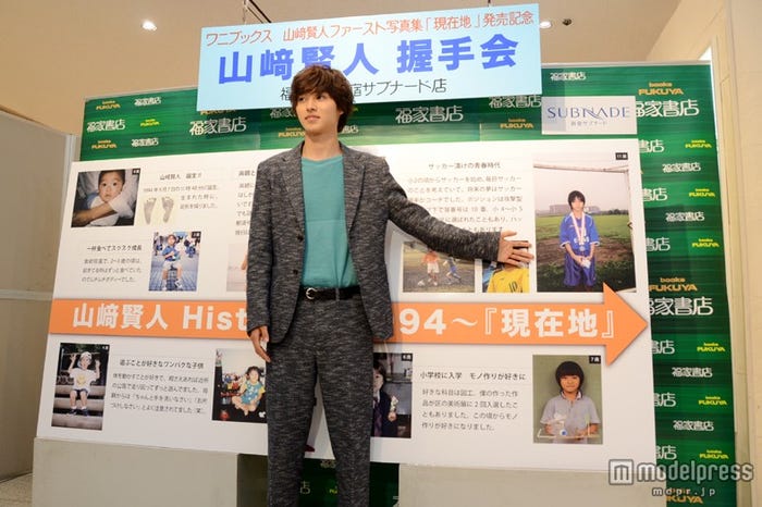 生い立ちを振り返る年表パネルをバックにポーズ