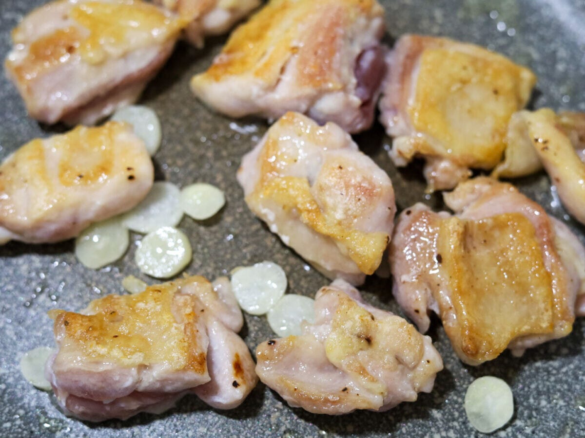 鶏もも肉とにんにくを焼く工程 フライパンでこんがり焼き色がついた鶏もも肉を裏返し、うす切りにんにくを加えて焼いている様子