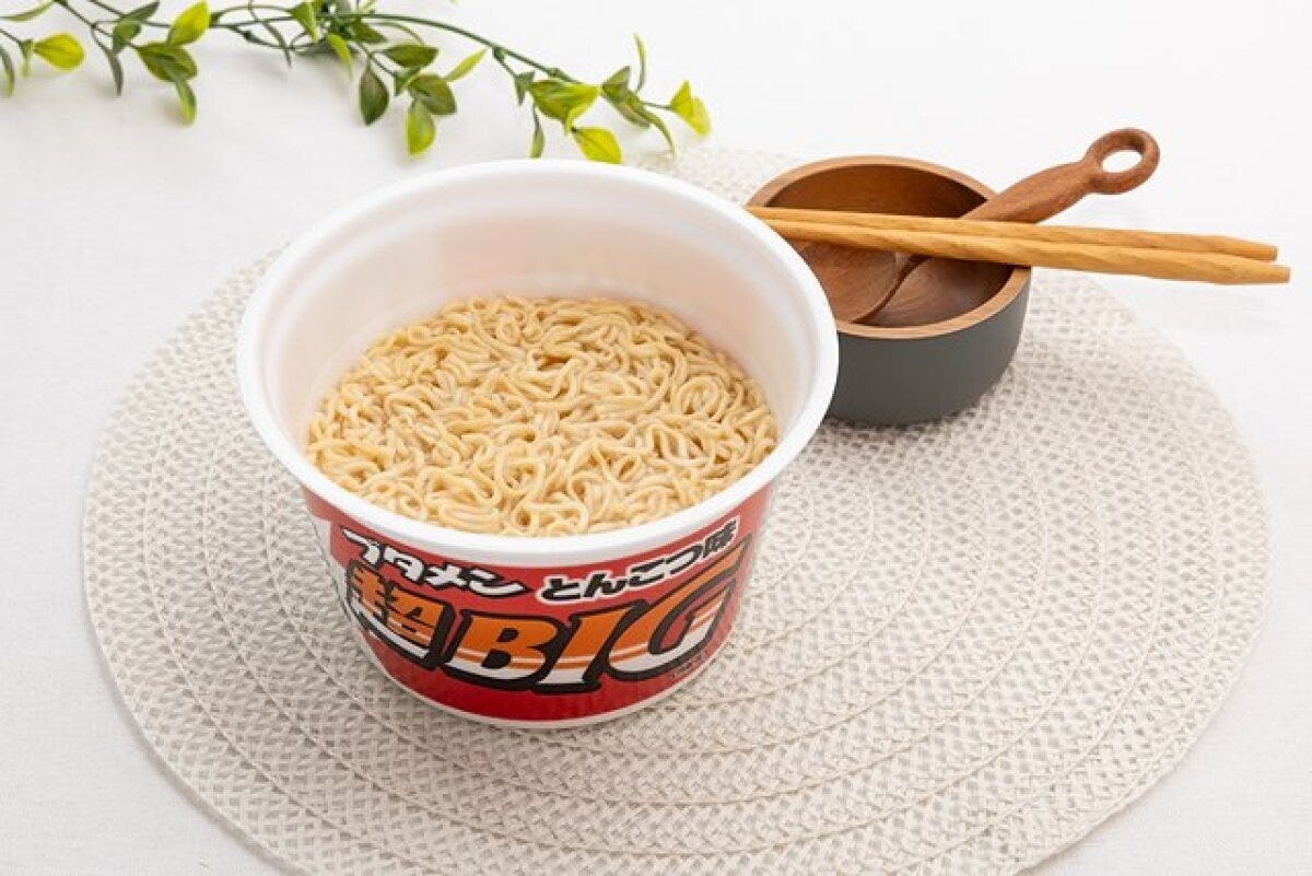 ブタメン超BIGの麺が見えるカップ麺が白い敷物の上に置かれている ブタメン超BIGの麺が見えるカップ麺が白い敷物の上に置かれている
