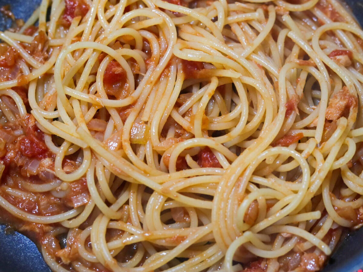 フライパンでトマトソースにスパゲッティを加えオリーブオイルをかけながらしっかり絡めている仕上げ工程写真