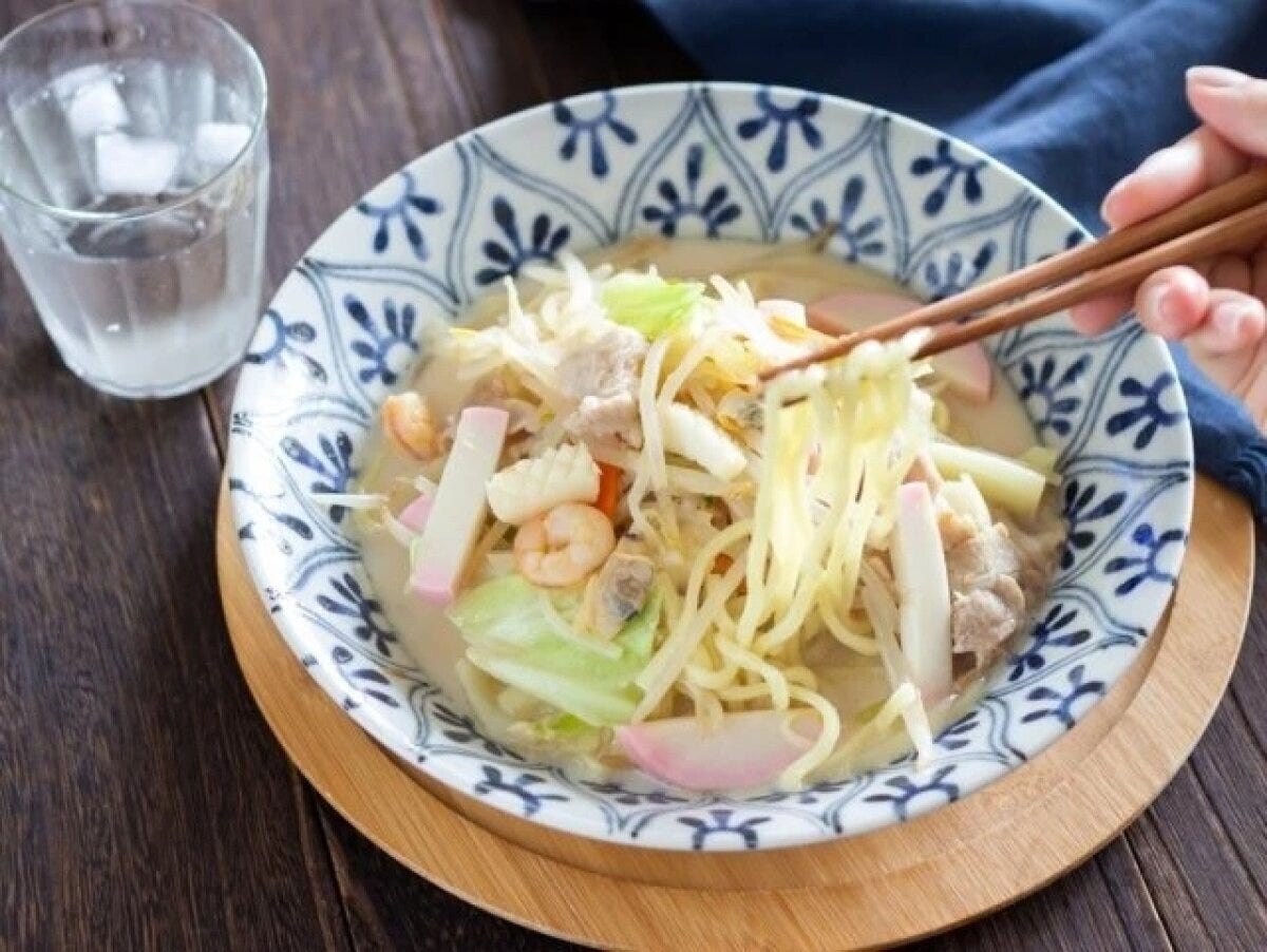 和柄の器に盛った具だくさん豆乳ちゃんぽんを箸で麺を持ち上げている写真。白濁スープに野菜・えび・かまぼこ・豚肉入り