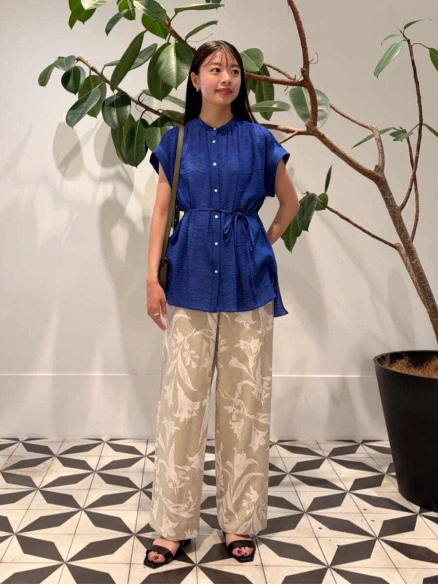 青いブラウスと花柄パンツの女性。黒サンダルを履き、観葉植物の横に立つ。