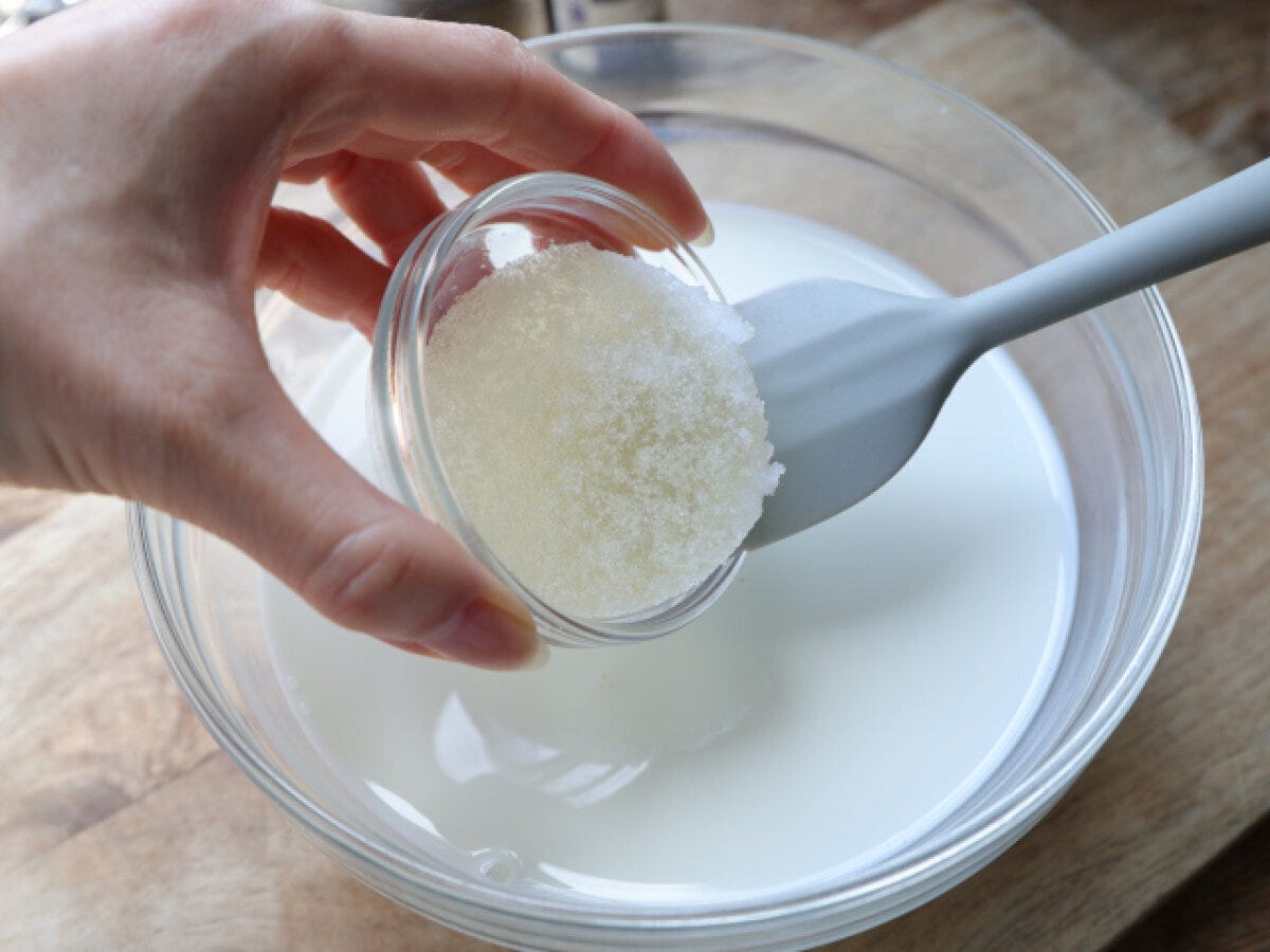 牛乳のボウルにふやかしたゼラチンを加えるところ 牛乳の入った大きなガラスのボウルに、ふやかしたゼラチンが入った小さなガラスの容器をシリコンヘラを添えて加えようとしている手元写真