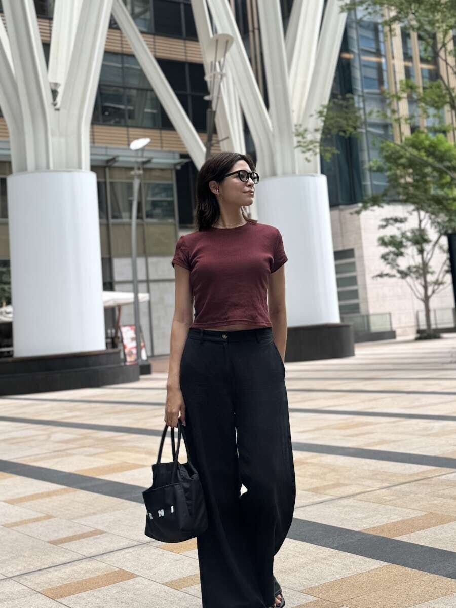 赤茶色のミニTと黒いワイドパンツを着用した女性が、街中でバッグを持って立っている
