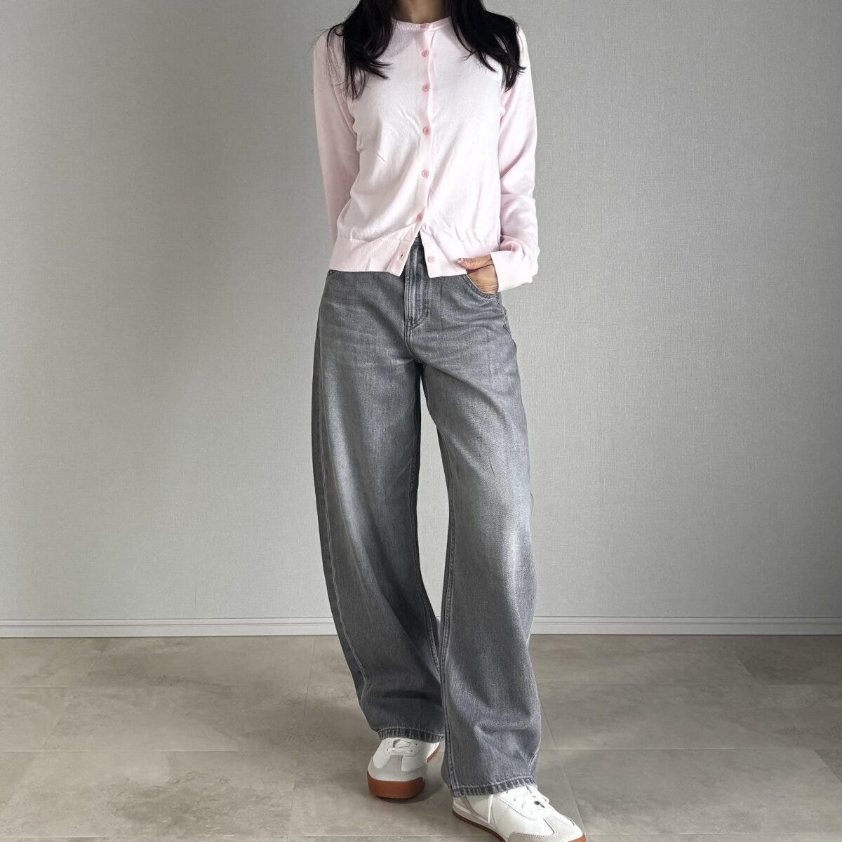 ピンクのカーディガンとグレーデニムを着用した女性が立つ