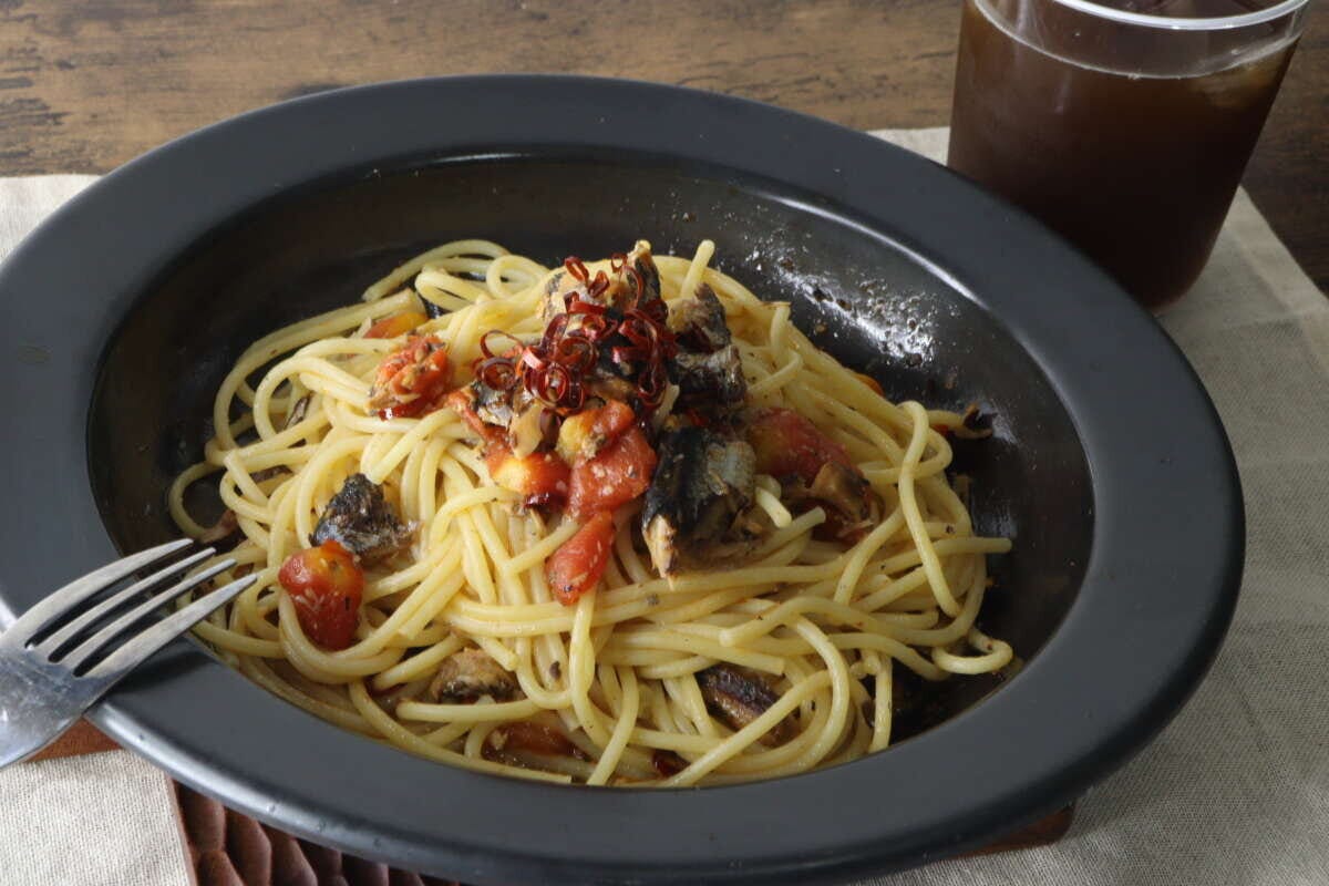 黒い皿に盛り付けられたいわしとミニトマトのオイルパスタ。奥にはグラスに入った飲み物が見える 黒い皿に盛り付けられたいわしとミニトマトのオイルパスタ。奥にはグラスに入った飲み物が見える