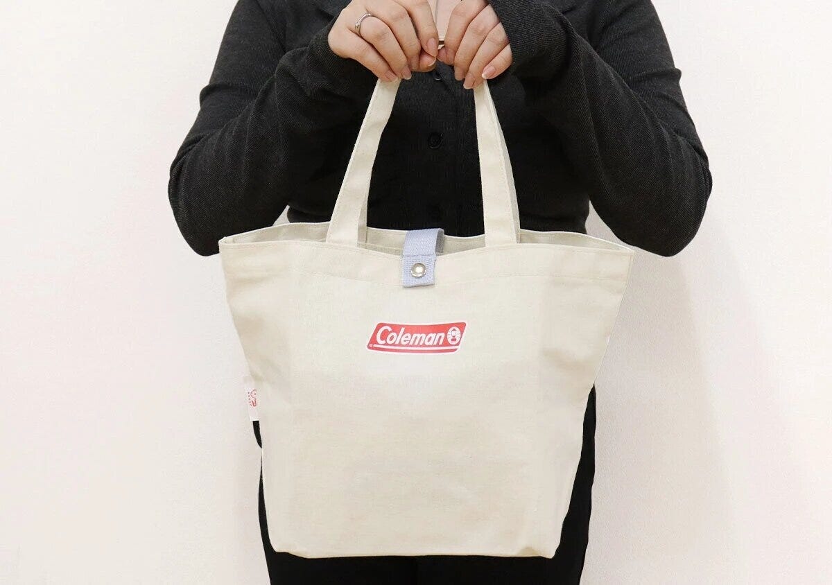 黒い服を着た女性がColemanロゴ入りナチュラルカラートートバッグを両手で持っている着用イメージ 黒い服を着た女性がColemanロゴ入りナチュラルカラートートバッグを両手で持っている着用イメージ