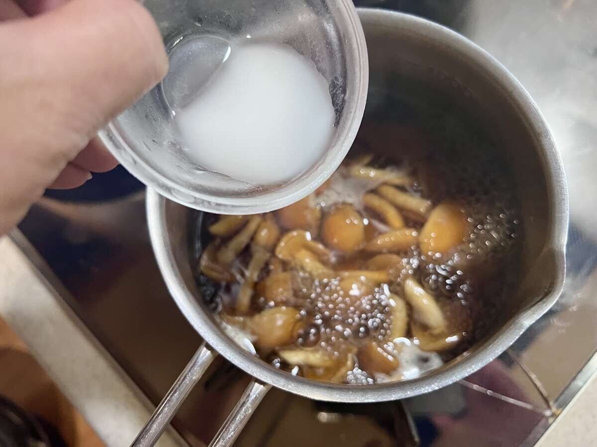 小鍋で煮込まれたなめこ入りのあんかけに、手で持った小皿から水溶き片栗粉が注ぎ入れられている