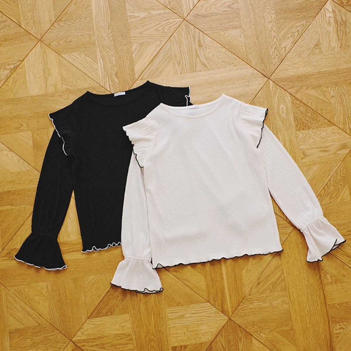 黒と白のフリルとメローデザインのプルオーバー2枚が木目調の床に並べられている 黒と白のフリルとメローデザインのプルオーバー2枚が木目調の床に並べられている