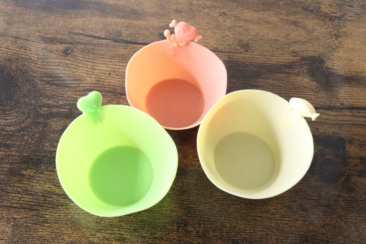 ピンク、緑、黄色のシリコンおかずカップ3つが木目のテーブルに並べられている ピンク、緑、黄色のシリコンおかずカップ3つが木目のテーブルに並べられている