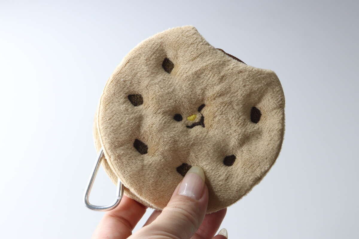 女性の指がクッキー型ポーチのカラビナ部分を持ち、ポーチの表面を見せている 女性の指がクッキー型ポーチのカラビナ部分を持ち、ポーチの表面を見せている