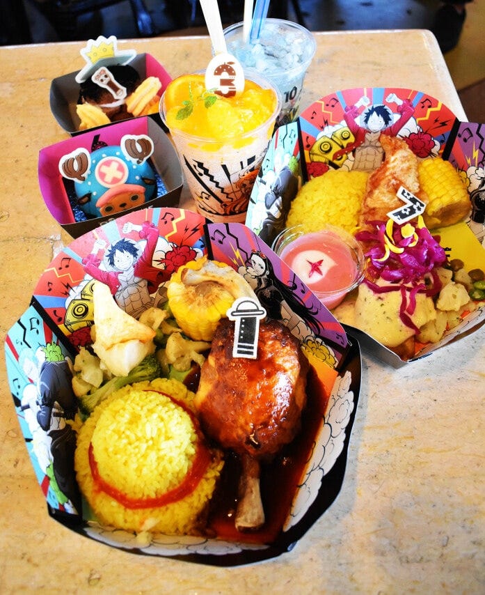 「ワンピース・サマー宴（フェス）・レストラン」のメニューは、右から時計回りに、ロビンのハナハナ・フィッシュフライプレート2000円、ルフィのでっかい骨付き肉メシ2100円、チョッパーのレモンムース850円、ブルックのマンゴー＆ココアシュークリーム850円。奥のドリンクは、ナミのオレンジフラッペ800円と操舵手ジンベエの海色ソーダ650円で「フォッシュル・フュエルズ」で楽しめます