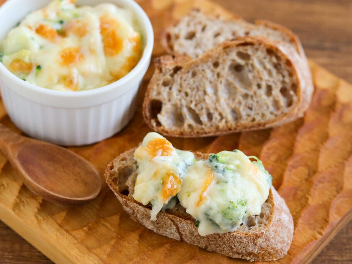 木のまな板の上にブロッコリーグラタンの皿とグラタンをのせたバゲット一切れが並んでいる様子 木のまな板の上にブロッコリーグラタンの皿とグラタンをのせたバゲット一切れが並んでいる様子