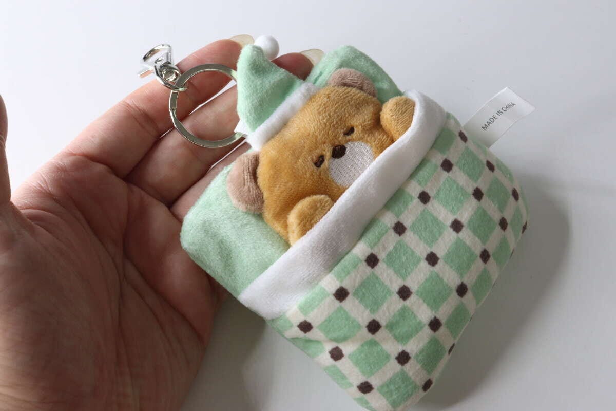 女性の手のひらに、緑の布団に入った茶色いくまのキーホルダーが乗っている 女性の手のひらに、緑の布団に入った茶色いくまのキーホルダーが乗っている