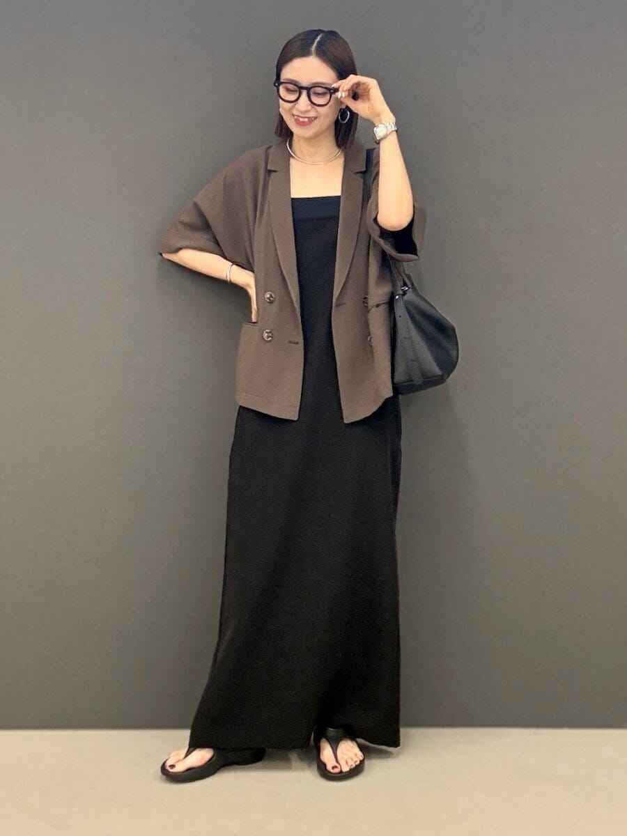 黒のキャミワンピースにブラウンの半袖ジャケットを羽織り、眼鏡をかけた女性が笑顔でポーズをとっている