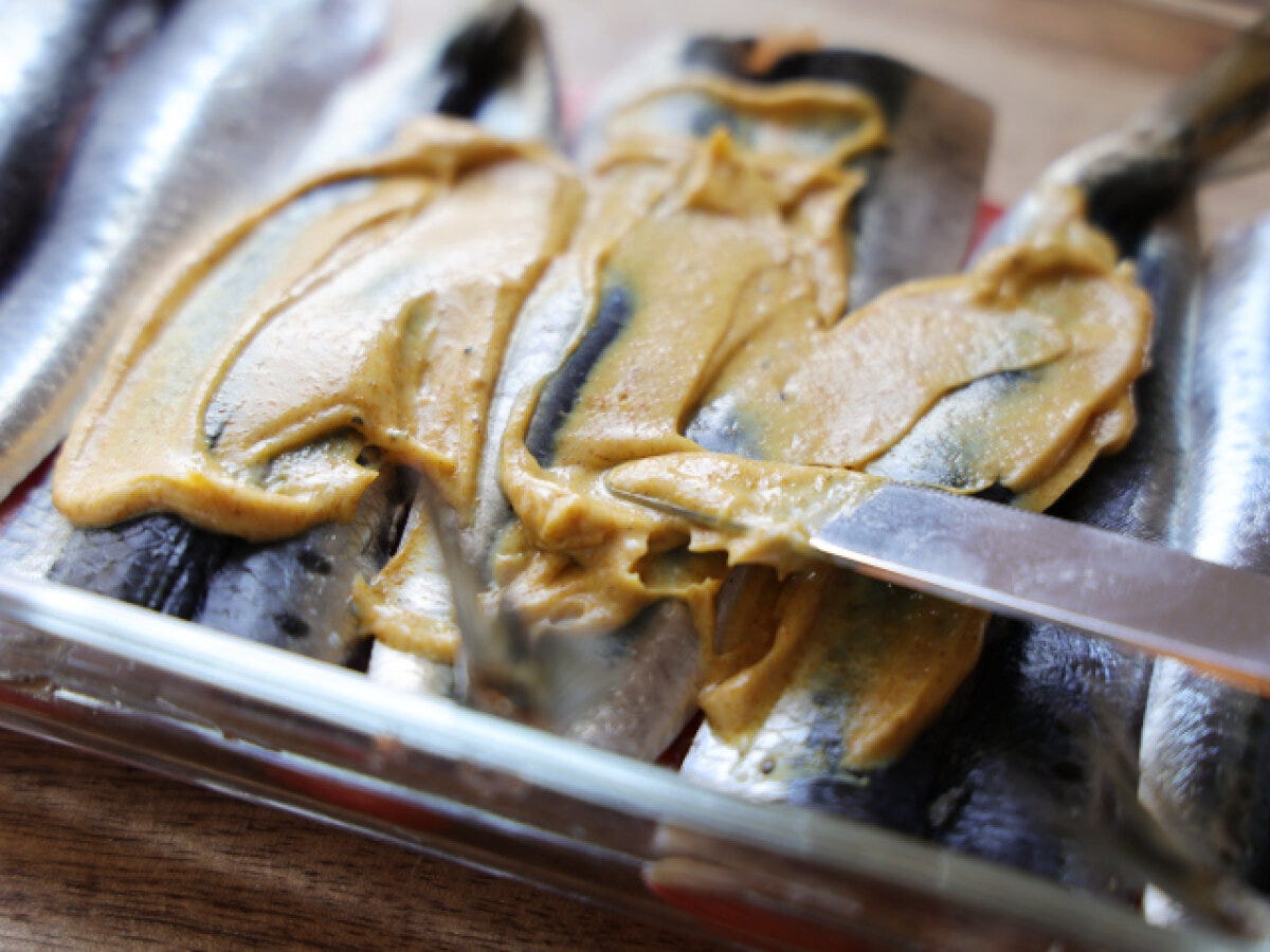 耐熱容器に並べられたいわしの皮目に、カレーマヨソースをスパチュラで塗っている様子 耐熱容器に並べられたいわしの皮目に、カレーマヨソースをスパチュラで塗っている様子