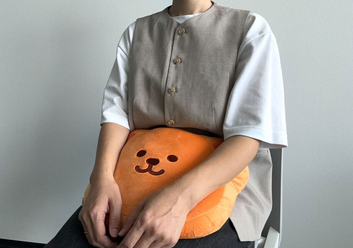 白いTシャツとベージュのベストを着た人が、ウィグルベアのもちもちクッションを膝の上に置いて座っている