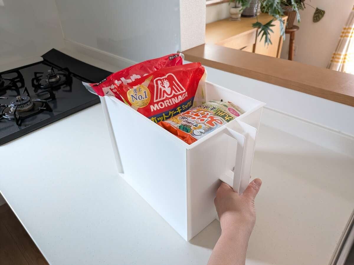 取っ手付き収納ボックスに食品ストックを立てて収納 白いキッチンカウンターの上で、取っ手付きの白い収納ボックスにホットケーキミックスやうどんなどの食品ストックを立てて収納し、手で取っ手を引いている写真