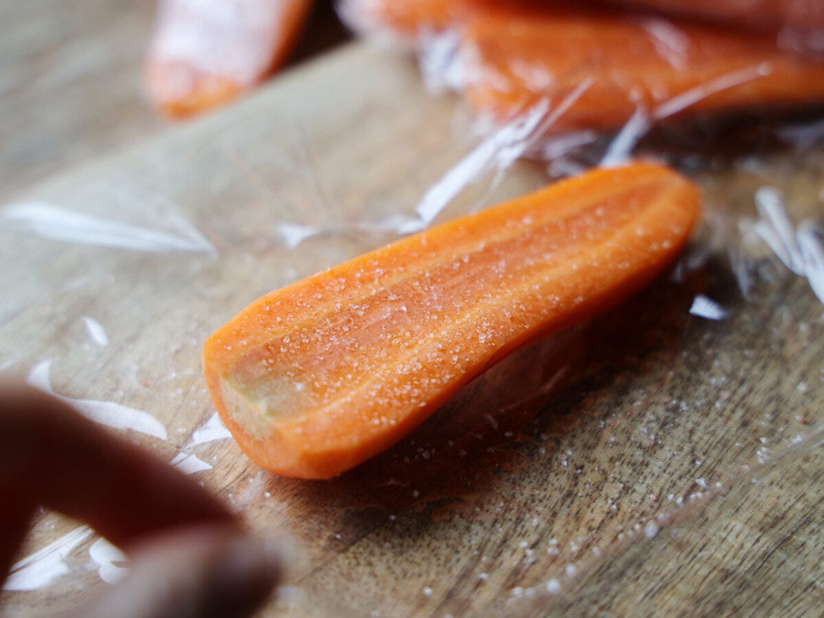 縦半分に切ったにんじんに塩を振った様子 ラップを敷いた木のまな板に縦半分に切ったにんじんを乗せ、塩を振った状態の写真