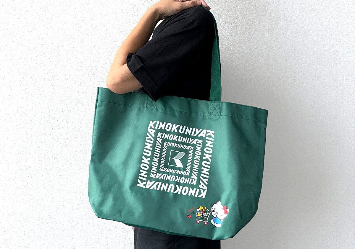 黒い服を着た人が緑色の紀ノ国屋×ハローキティお買い物バッグを肩にかけている