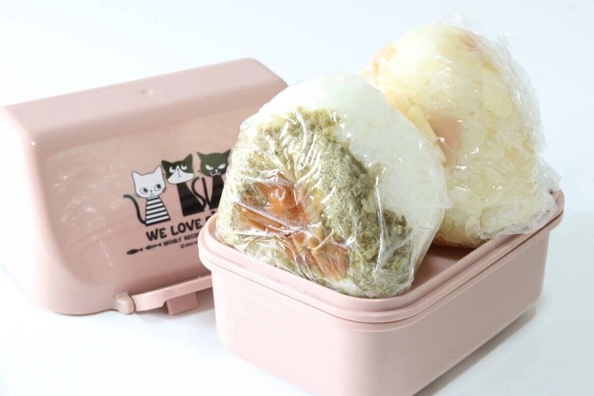 ピンク色の猫柄弁当箱の隣にラップおにぎり2個が置かれている