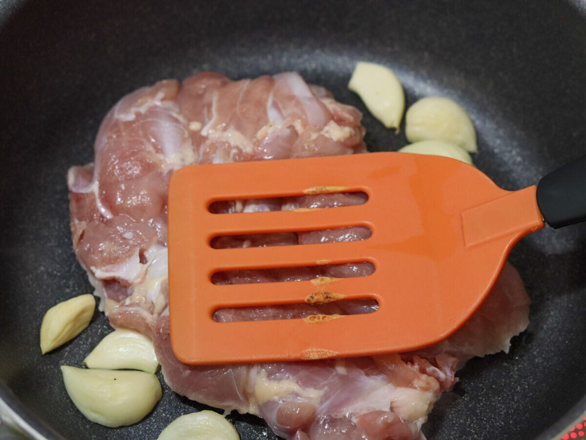 オレンジ色のヘラで鶏もも肉を押さえながらフライパンで焼いている様子