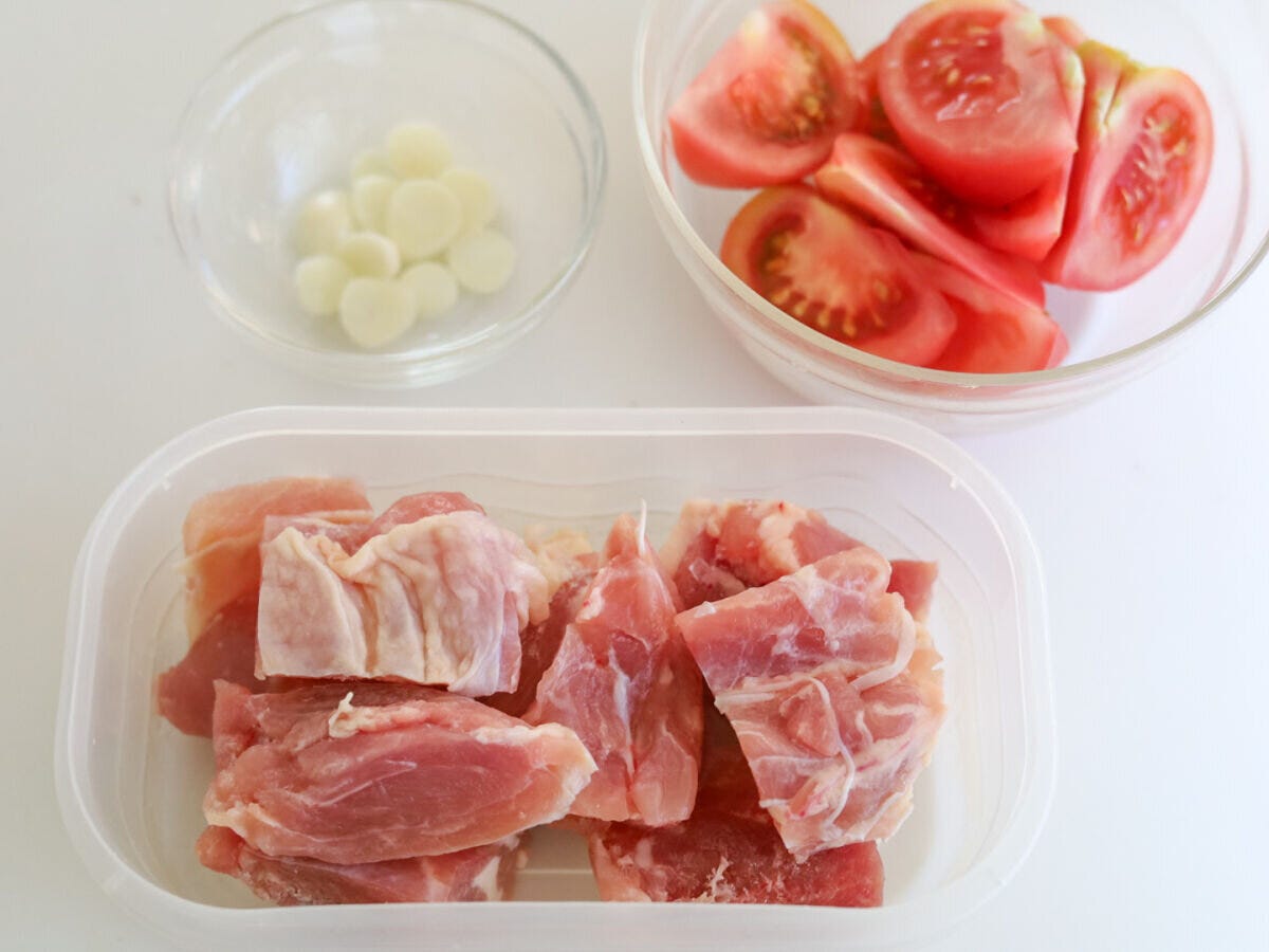 鶏もも肉・トマト・にんにくの下ごしらえ 白いテーブルに並んだ、うす切りにしたにんにく・くし切りにしたトマト・一口大に切った生の鶏もも肉の下準備の様子