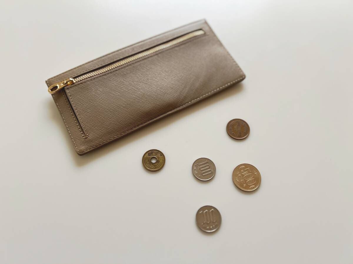 白いテーブルの上に置かれたブロンズ色の薄型長財布と、5円・10円・100円などの硬貨が数枚並んでいる