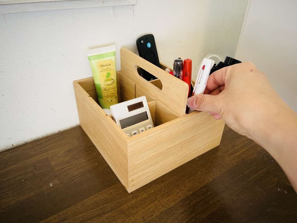 仕切り付き木製ボックスにペン・リモコン・電卓を立てたワンアクション収納 木製の仕切り付き収納ボックスに立てて入れたハンドクリーム・リモコン・電卓・ペンから手でペンを取り出している様子
