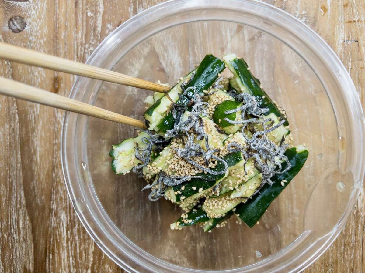 透明なボウルの中でちぎったきゅうりと塩昆布、ごまを箸で混ぜている俯瞰