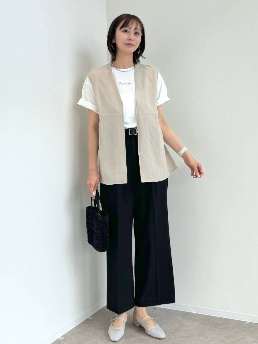 ベージュジレと白T、黒パンツを着た女性が壁を背景に立つ