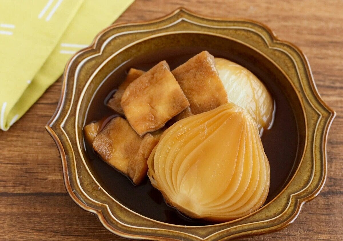 半分に切った新玉ねぎと四角く切った油揚げを、濃い茶色のだし汁で煮たスープ煮を、茶色い花型の器に盛った写真