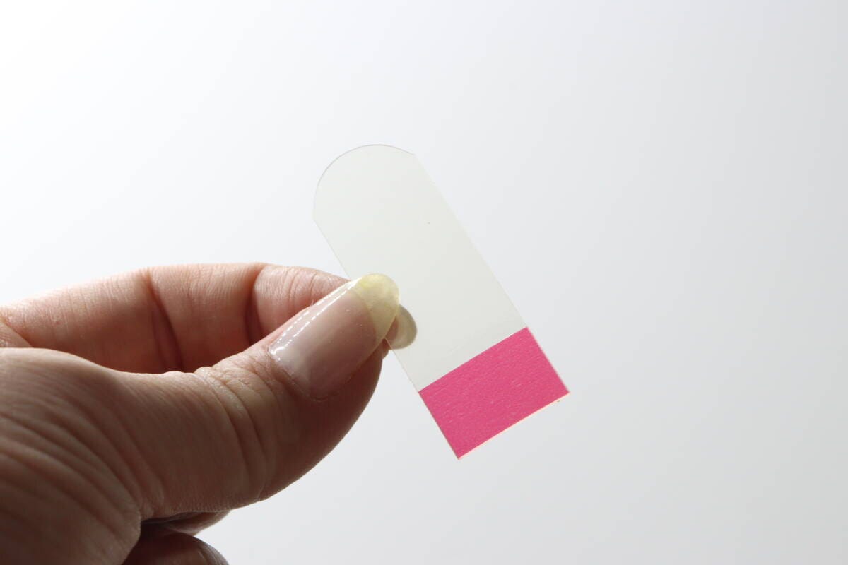 指で透明な伸びるテープを1枚持ち、赤い保護フィルムが見えている
